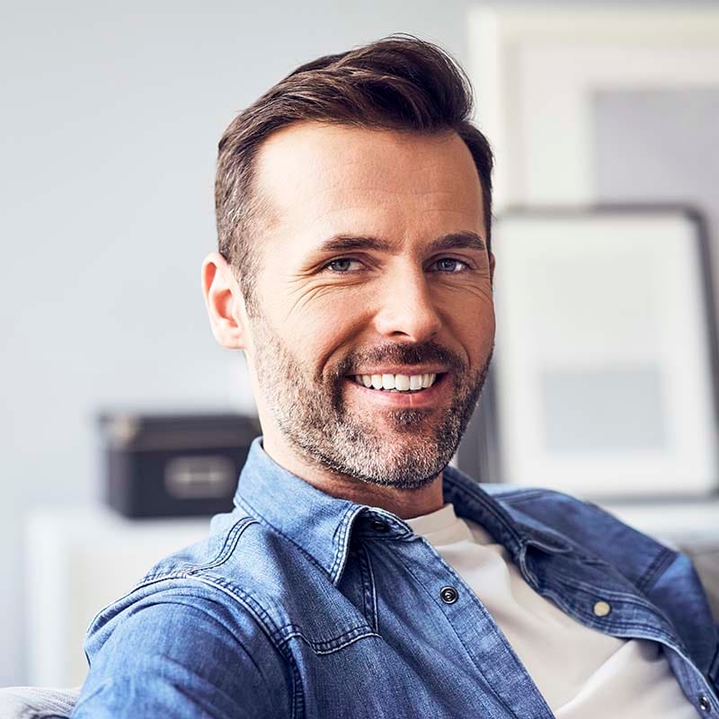 Man sitting on sofa smiling into camera wearing blue denim snap-up collared shirt layered over white casual-dress t-shirt