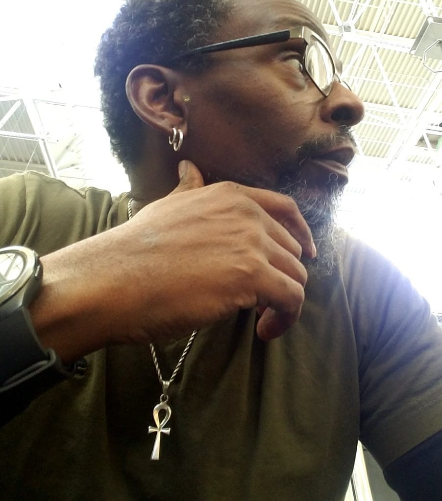 Image of BLACK man with curly short hair glasses silver earrings sports watch ankh necklace green shirt hand on chin looking to away from camera to the right as you face the screen