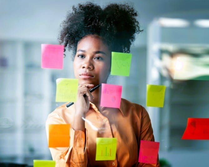 Thoughtful ABiAn™ young woman strategizing at glass board considering the options on post-its sticking to the glass.