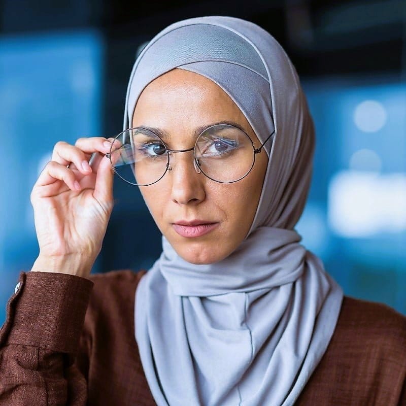 Muslim Euro woman with gray hijab peaking over the top of larger rounded glasses, wearing a brown shirt with button sleeves.