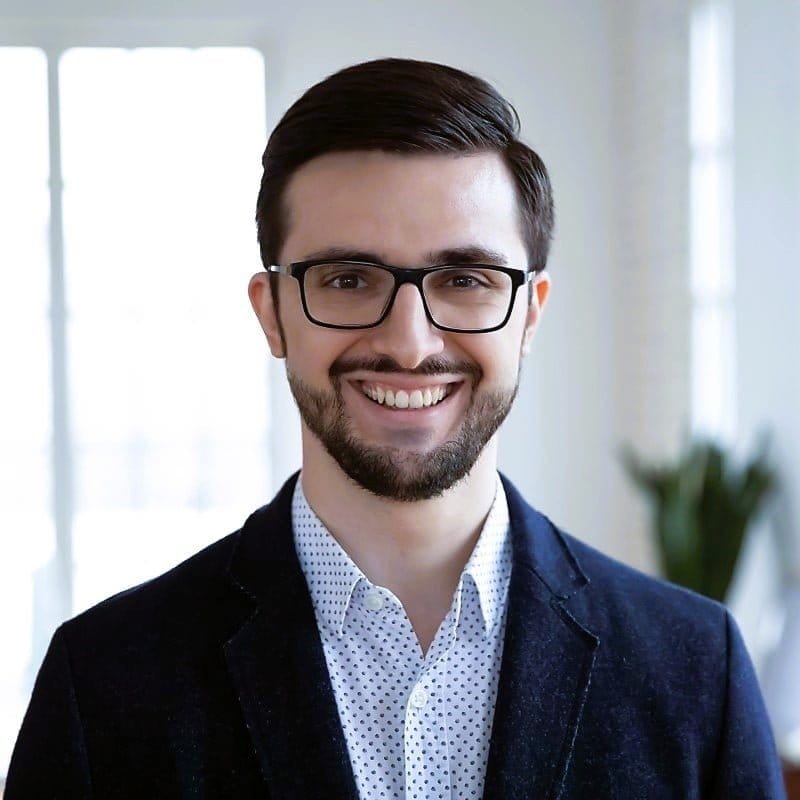 Euro youngish businessman with glasses, stubble beard, moustache, navy blue blazer, button down, light blue shirt, cheery smile, green plant and ceiling to floor window in blurred background