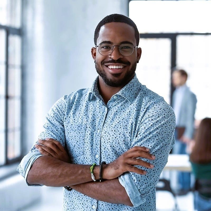 ABiAn young male, casual short beard and moustache entrepreneur, smiling, arms relaxingly folded in front, sleeves rolled to elbow blue dot patterned button shirt. Earthy bracelets are visible on both wrists, two on one. He is standing in the foreground of a tabled room, people behind him sitting and a person standing with a portfolio in his hand.