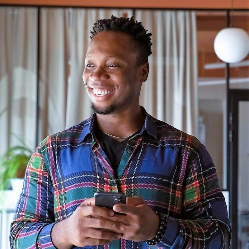 ABiAn young, business male, scruffy short beard, no moustache, sporting trendy short spike hair twists style, casually dressed in blue, orange, green plaid button shirt, also wearing warm cheery smile and holding black smartphone in both hands held chest high. He is standing in his restaurant.