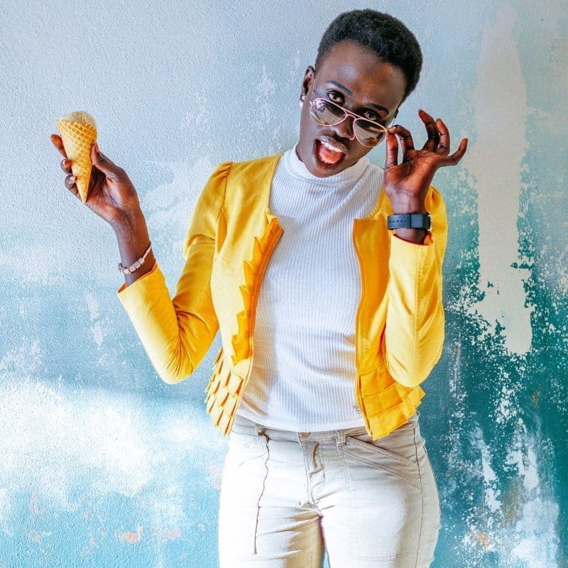 ABiAn unidentifiable gender, sassy, yellow jacket layered over white top, fitted beige pants, peeking over sunglasses tipped down by left hand.