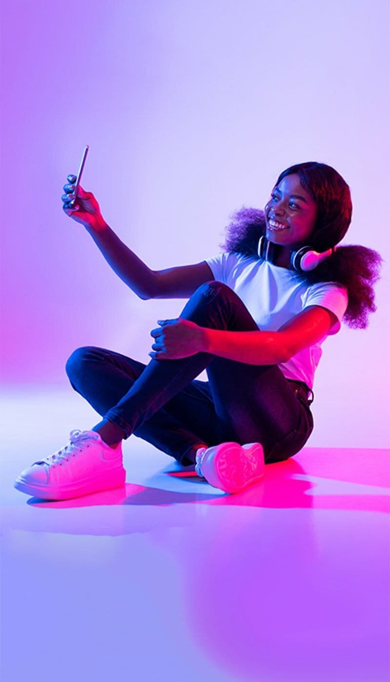 African-American (ABiAN) young female vlogger sitting on the floor in a room washed with hures of purple and pink. She has headphones draped around her neck while she smiles into smartphone taking selfie.